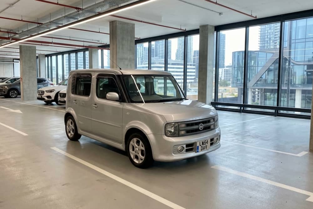 Silver Nissan Cube parked in a car park, a compact Japanese import known for its boxy design.